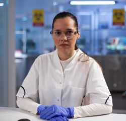 A woman in lab coat and gloves sitting at a table.